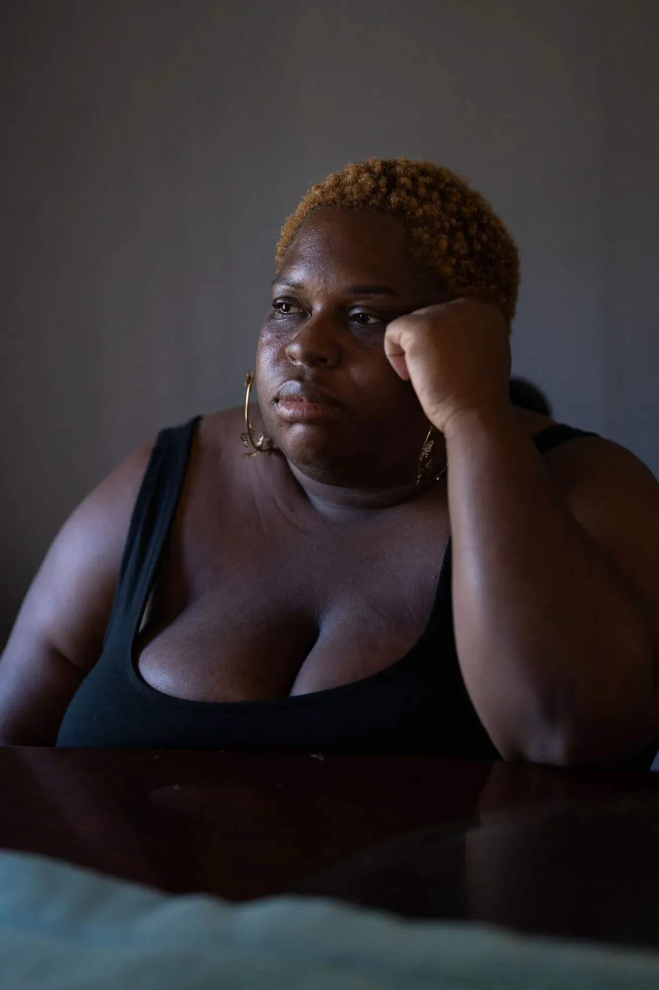 امرأة ذات بشرة داكنة تجلس في غرفة مضاءة بشكل خافت، تعبر عن مشاعر القلق والتفكير العميق، مما يعكس التوتر المرتبط بالقرارات الطبية أثناء الحمل.