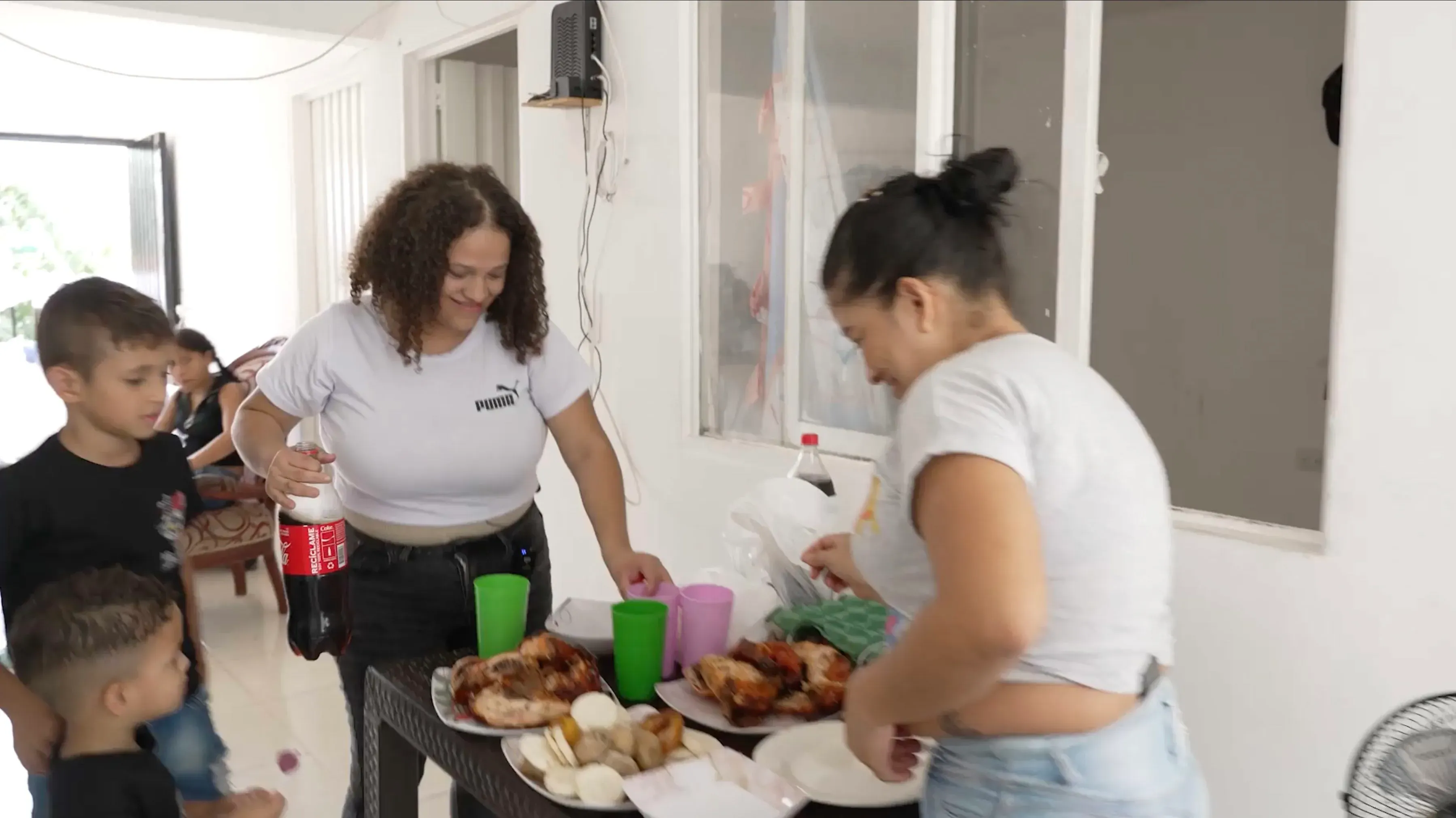 امرأتان مع أطفال في مطبخ، واحدة تحضر الطعام والأخرى تحمل زجاجة مشروب، تعكس الحياة اليومية للمهاجرين في كولومبيا.