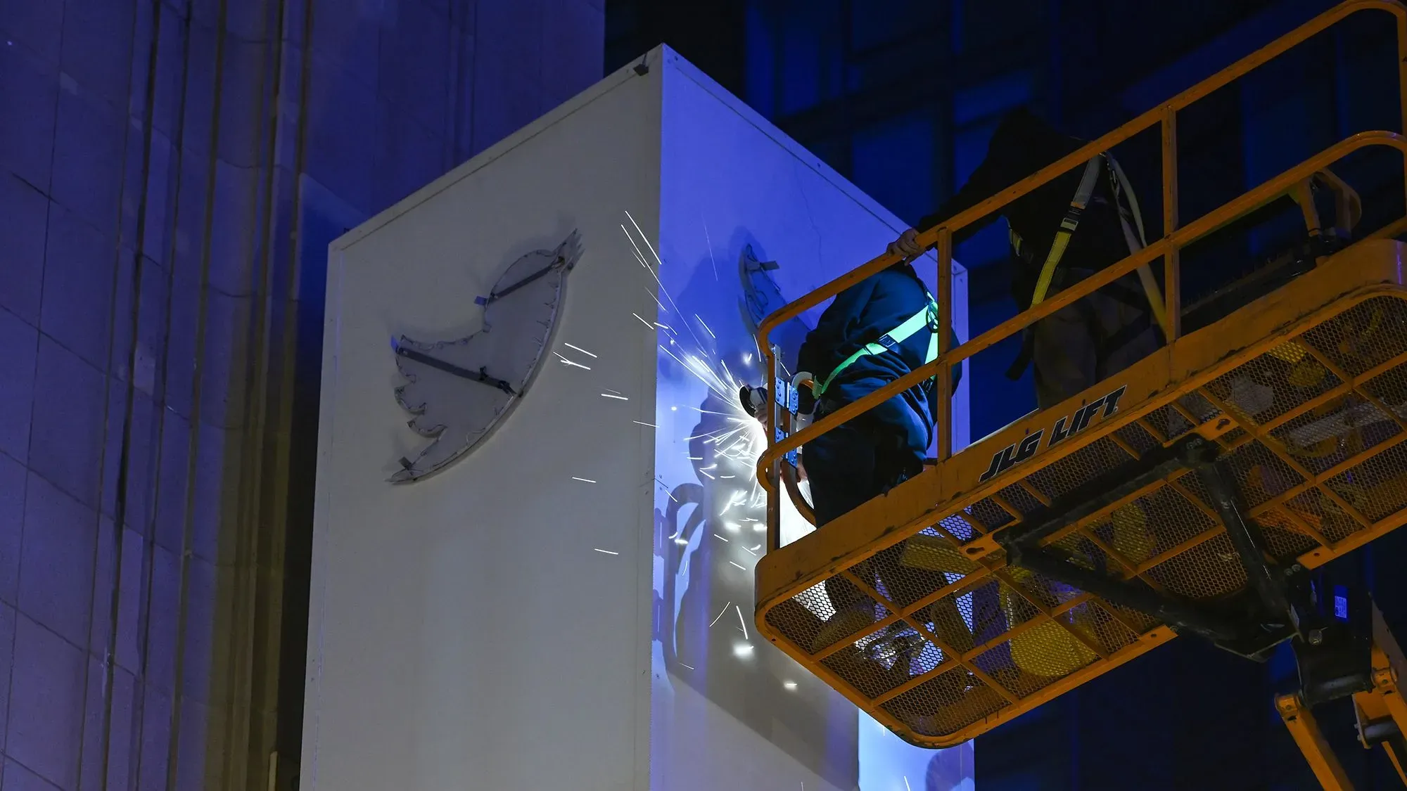 عمال يقومون بإزالة شعار تويتر من المبنى باستخدام معدات الرفع، مع إضاءة زرقاء في الخلفية، مما يعكس التحولات في الشركة.