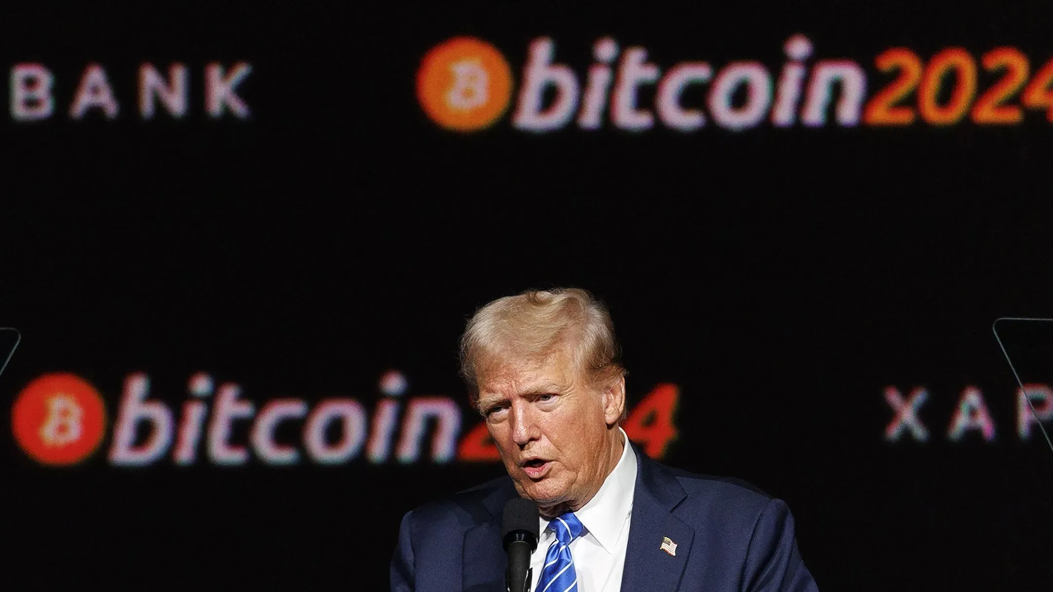 دونالد ترامب يتحدث في حدث مرتبط بعملة البيتكوين، مع شعار "bitcoin 2024" في الخلفية، مشيرًا إلى اهتمامه بالعملات الرقمية.