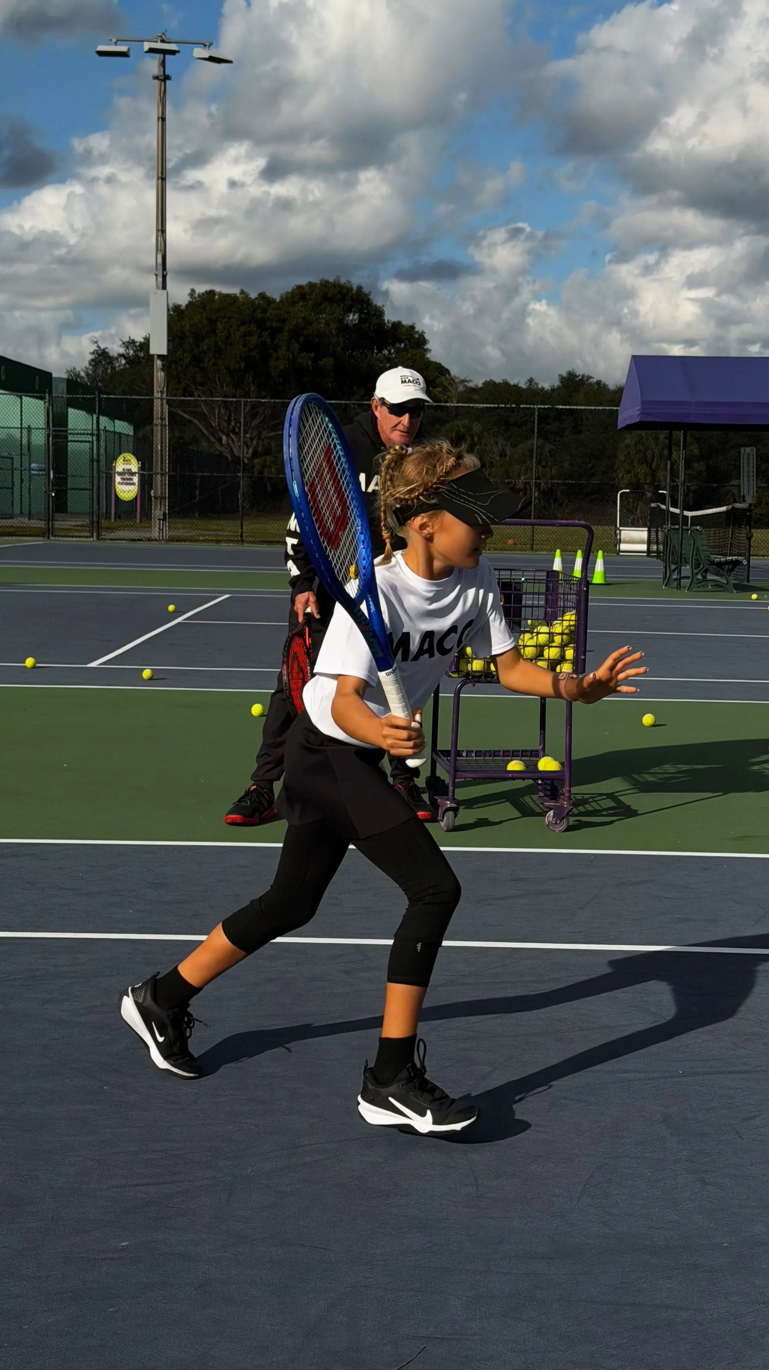 طفلة صغيرة تمارس التنس تحت إشراف مدربها، تتدرب بجد على ملعب، مع وجود كرات تنس حولها، تعكس شغفها للرياضة.