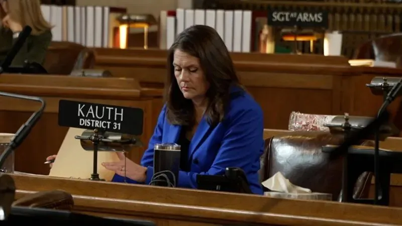 امرأة ترتدي بدلة زرقاء تجلس في قاعة مجلس تشريعي، تركز على الأوراق أمامها. تعكس الصورة الأجواء السياسية في نبراسكا وسط قلق المهاجرين.