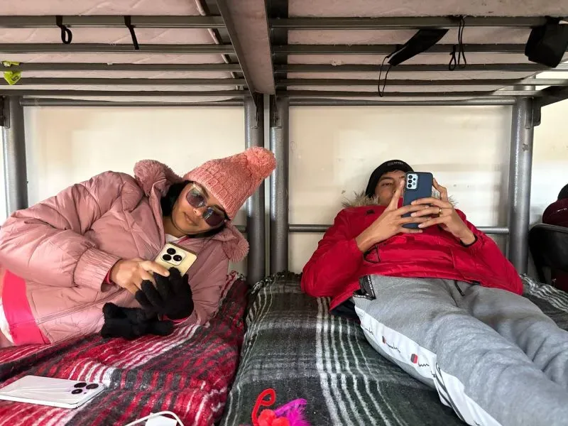 امرأة ترتدي معطفًا ورديًا وقبعة جليدية، تجلس على سرير في ملجأ، وتستخدم هاتفها. بجوارها، شاب يرتدي سترة حمراء، مشغول بهاتفه أيضًا، في أجواء هادئة تعكس حياة المهاجرين.