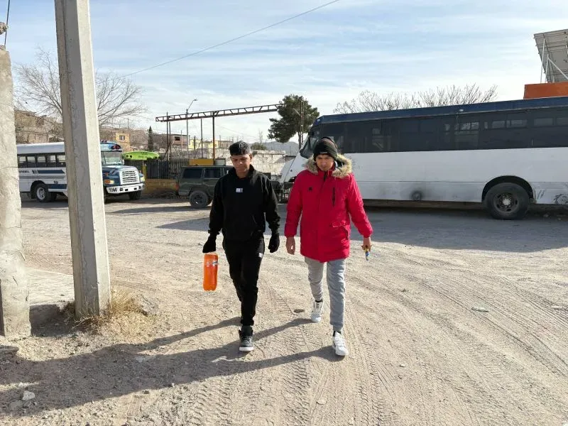 شابان يسيران في منطقة مفتوحة بالقرب من حافلات، أحدهما يرتدي معطفًا أحمر والآخر ملابس داكنة، في أجواء شتوية باردة.