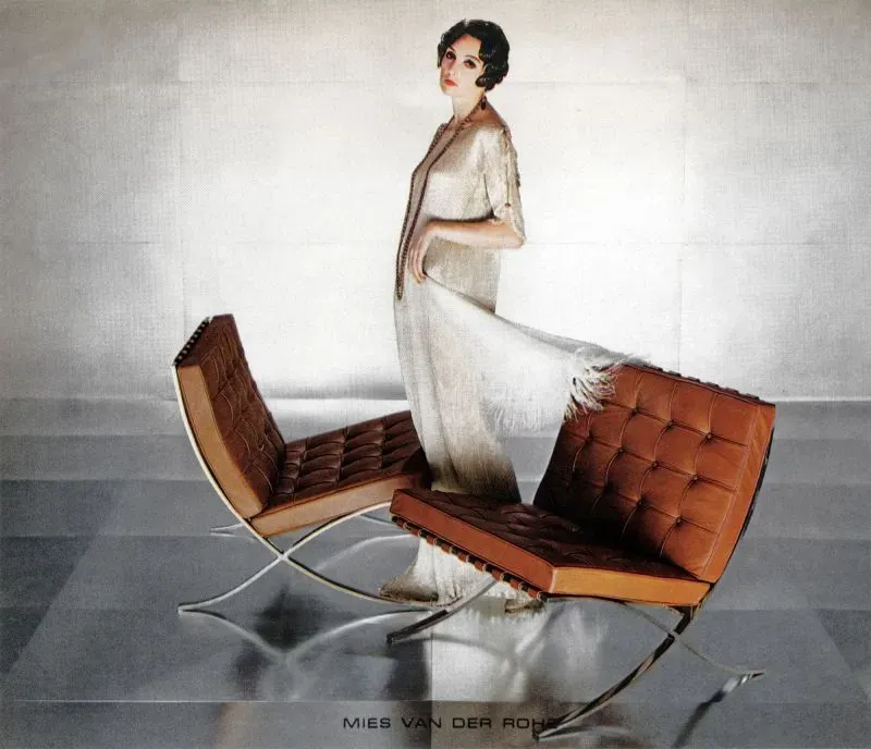 امرأة ترتدي فستانًا أنيقًا تقف بجانب كرسي برشلونة الشهير بتصميمه العصري، مع تفاصيل جلدية وإطار معدني، يجسد روح الحداثة.