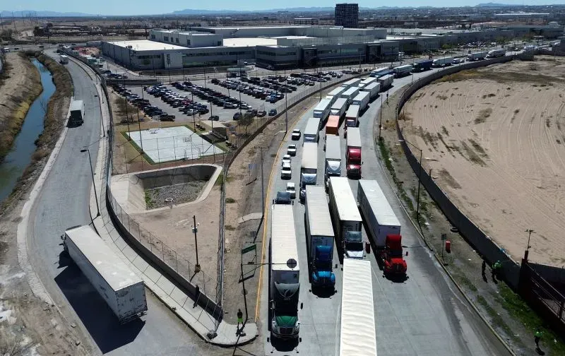 طوابير طويلة من الشاحنات عند نقطة عبور حدودية، مع مباني خلفية، تعكس حالة عدم اليقين الاقتصادي وتأثير التعريفات الجمركية.