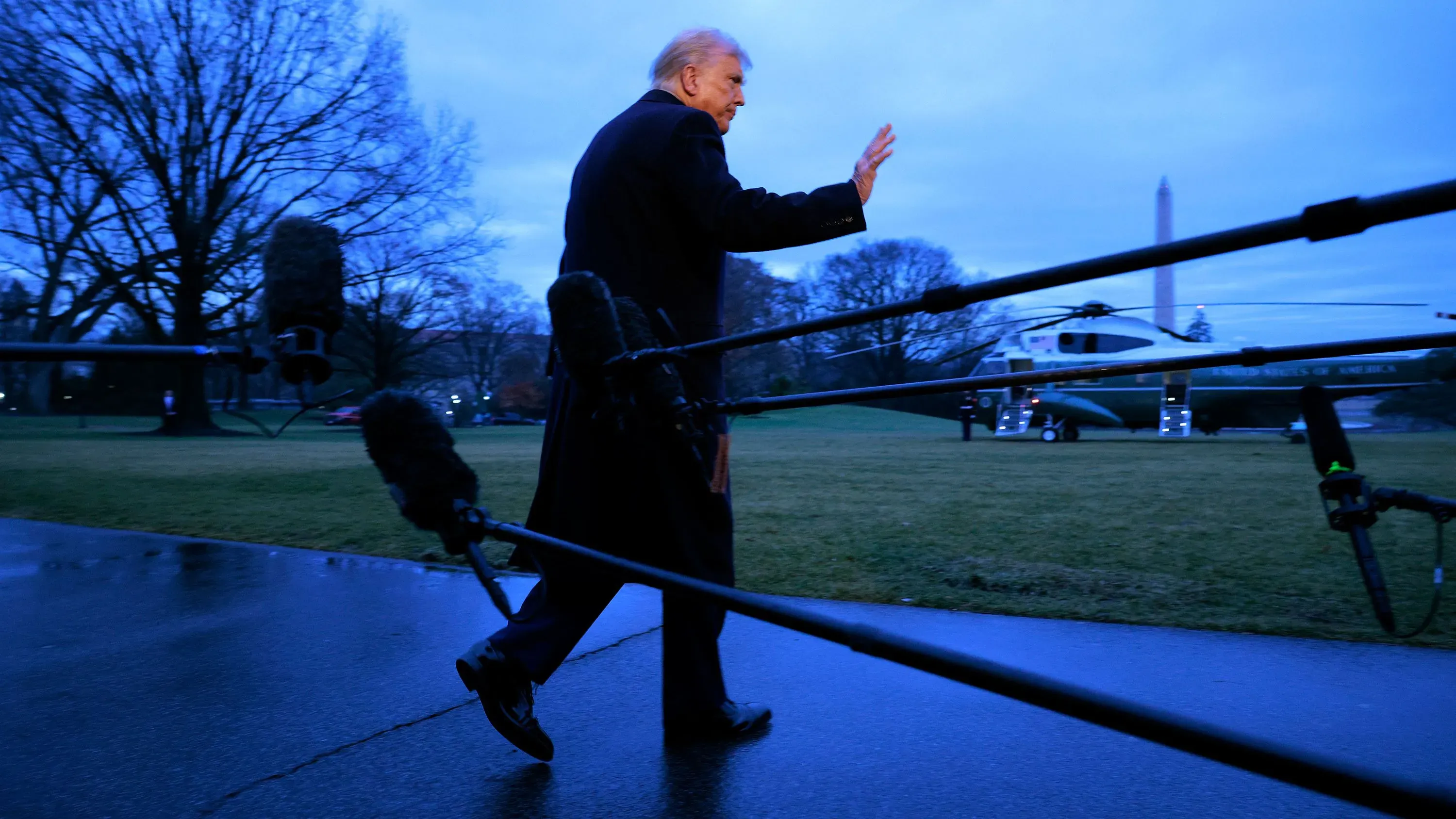 رجل يسير في حديقة البيت الأبيض، يلوح بيده، مع ظهور طائرة هليكوبتر ومبنى واشنطن في الخلفية، مما يعكس حالة عدم اليقين السياسي.