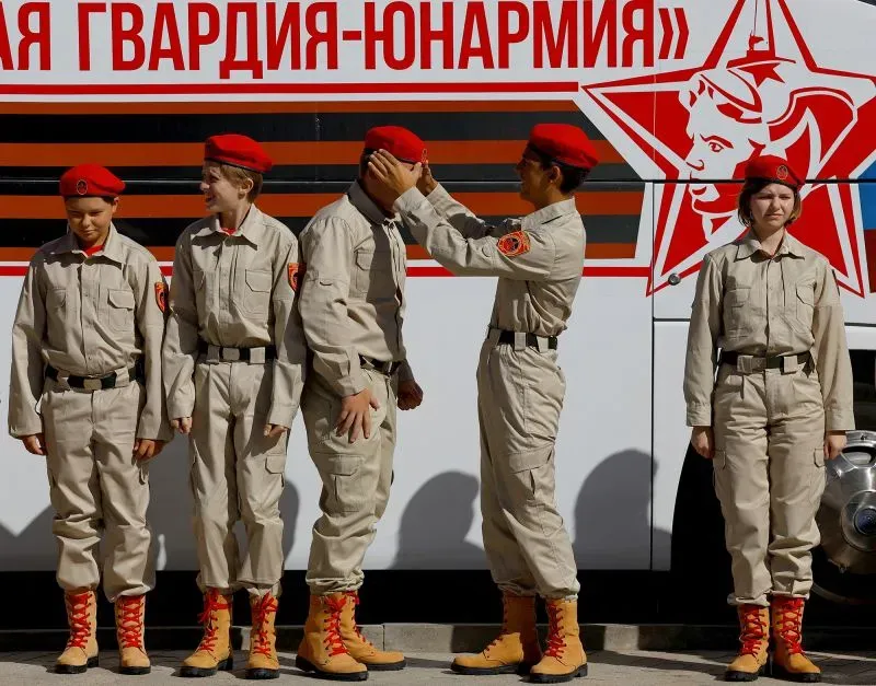 مجموعة من الشباب يرتدون زيًا عسكريًا، مع قبعات حمراء، يقفون أمام حافلة تحمل رموزًا عسكرية، مما يعكس تأثير الاحتلال الروسي في أوكرانيا.