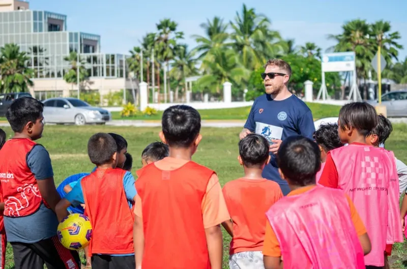 مدرب يتحدث إلى مجموعة من الأطفال يرتدون قمصاناً ملونة أثناء تدريب كرة القدم في جزر مارشال، وسط أجواء مشمسة وخلفية طبيعية جميلة.