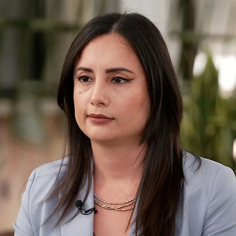 امرأة ذات شعر بني طويل ترتدي سترة رمادية، تبدو متفكرة. تعكس ملامح وجهها القلق بشأن قضايا الهجرة.