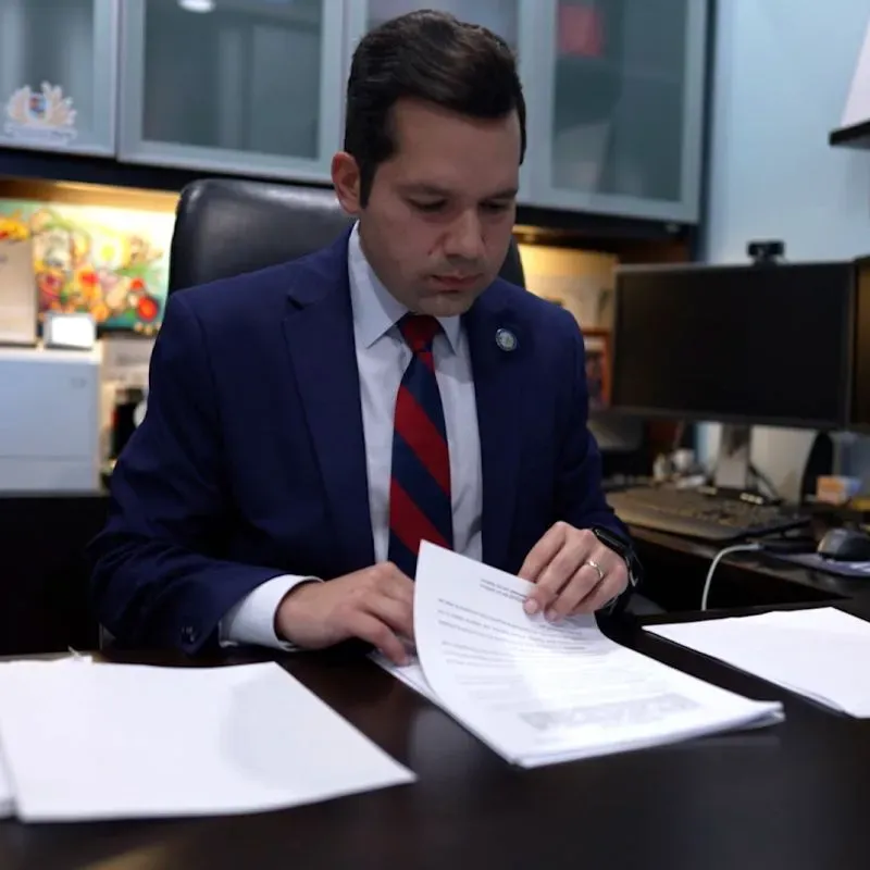 رجل يرتدي بدلة رسمية يجلس على مكتب، يراجع مستندات مهمة، مع خلفية تحتوي على أجهزة كمبيوتر ومطبوعات ملونة.