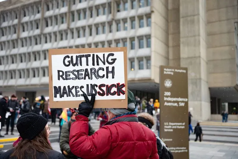 لافتة في مظاهرة تحمل عبارة "تخفيض الأبحاث يجعلنا مرضى" أمام مبنى حكومي، تعبيرًا عن الاحتجاج ضد تقليص ميزانية الأبحاث.
