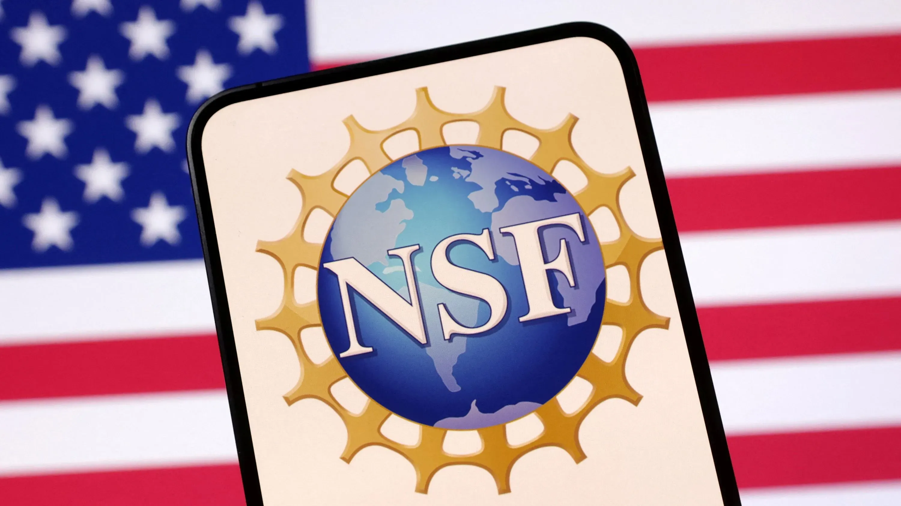 شعار المؤسسة الوطنية للعلوم (NSF) على شاشة هاتف محمول، مع العلم الأمريكي في الخلفية، يعكس الجدل حول سياسات التمويل البحثي.