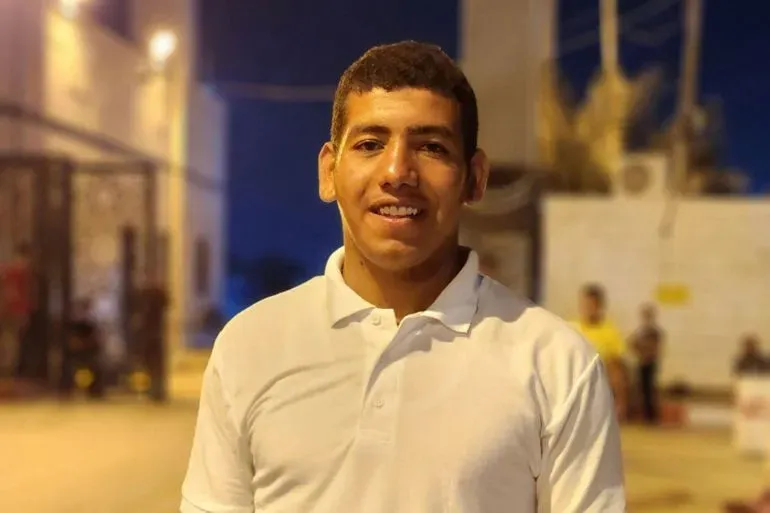 شاب مبتسم يرتدي قميصًا أبيض، يقف في موقع توزيع المساعدات في غزة، مع خلفية مضاءة، تعكس الوضع الإنساني الصعب.