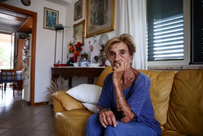 امرأة مسنّة تجلس في صالة منزلها، تعبر عن قلقها بشأن مشروع جسر مضيق ميسينا وتأثيره على مجتمعها.