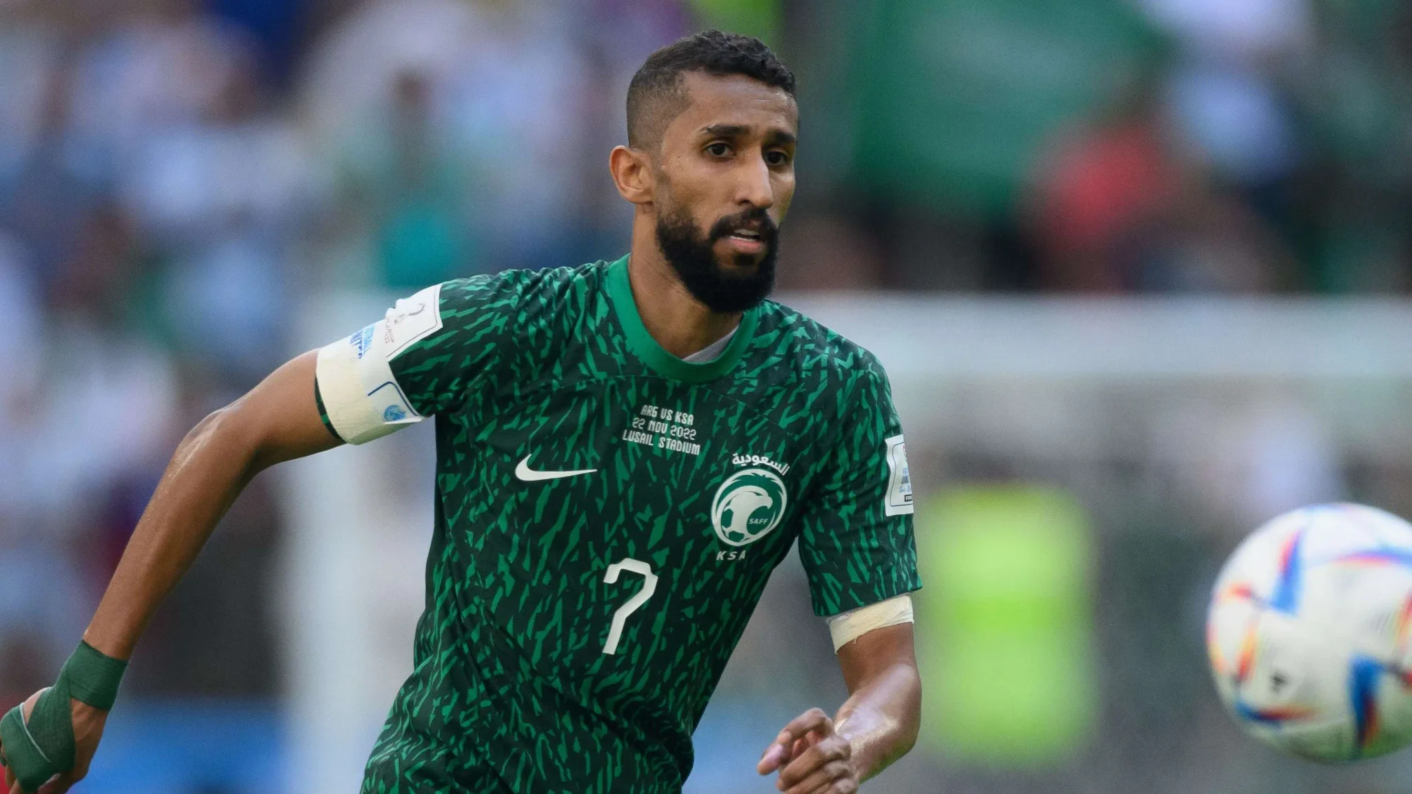سلمان الفرج قائد منتخب السعودية السابق يرتدي الزي الأخضر يركض خلف الكرة خلال مباراة دولية، مع خلفية جماهيرية ضبابية.