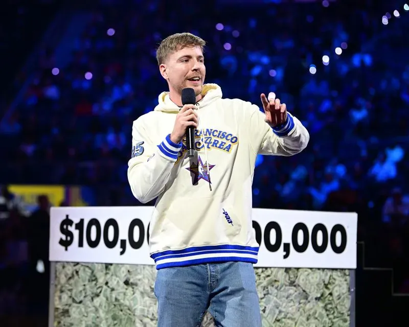 شخص يتحدث على المسرح أمام جمهور، يرتدي سترة بيضاء مع شعار، ويشير نحو لافتة مكتوب عليها "$100,000"، في حدث يتعلق بتطبيق TikTok.