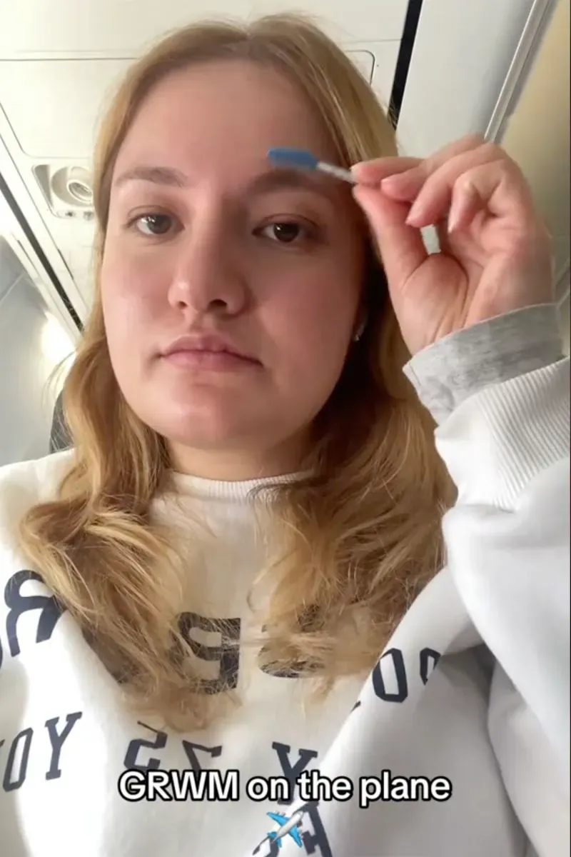 امرأة شابة في الطائرة، تستخدم فرشاة لتصفيف حواجبها، مرتدية سترة مريحة، تعبر عن تجربة العناية الذاتية أثناء الرحلة.