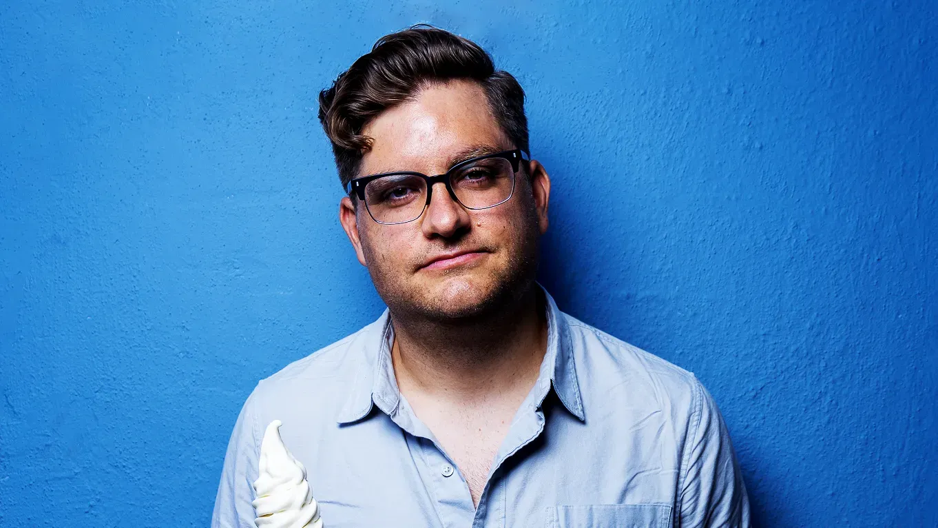 أليكس فالكوني، ممثل كوميدي معروف، يحمل آيس كريم، أمام خلفية زرقاء، يتحدث عن فنون الاحتيال وتقبل الجمهور لها عبر وسائل التواصل.