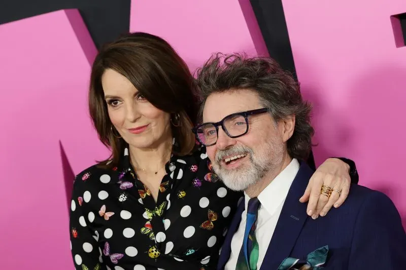 تينا فاي وجيف ريتشموند يتبسمان معًا في حدث عام، بينما يرتديان ملابس أنيقة، مع خلفية وردية تضيف لمسة مرحة.