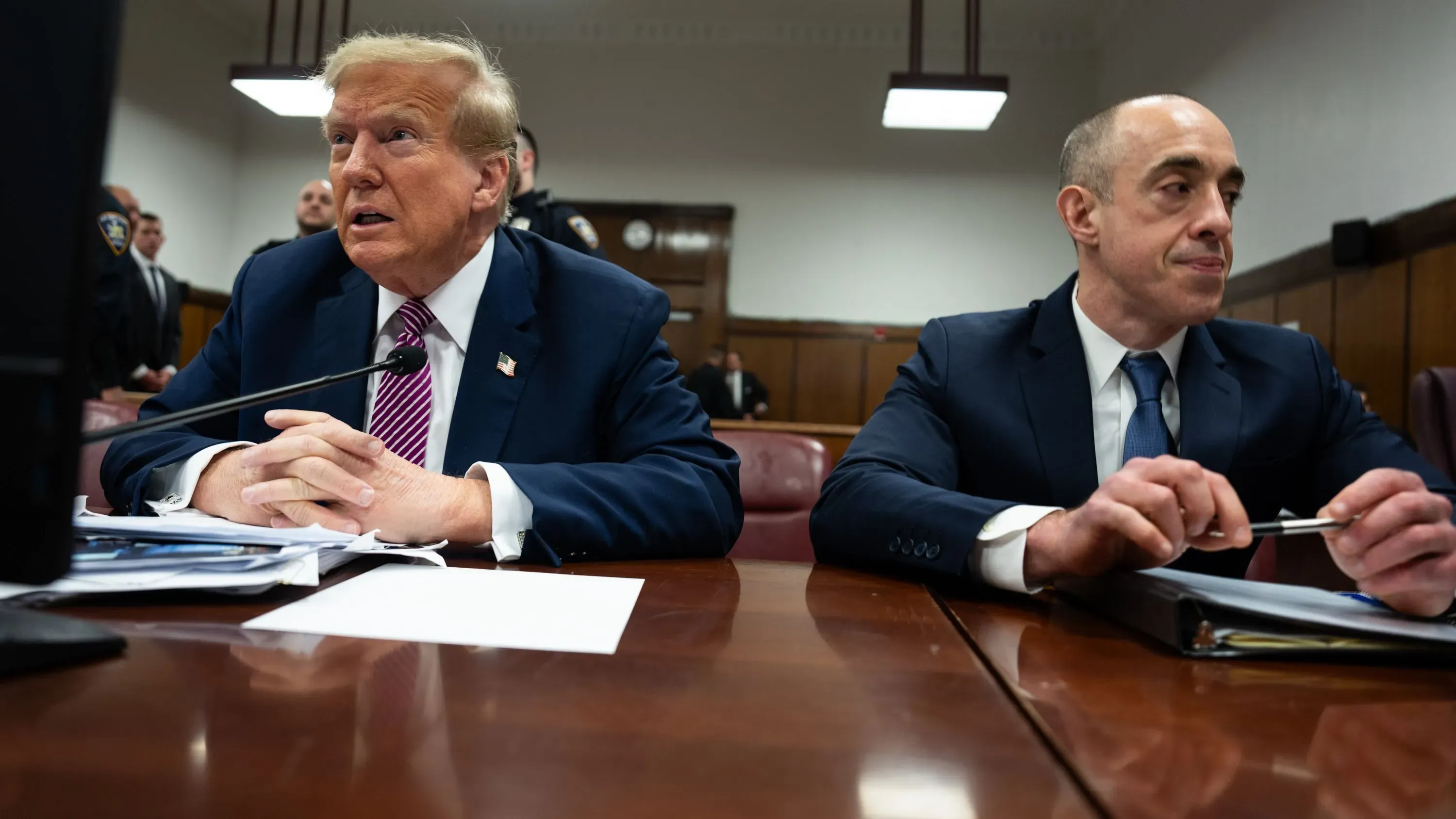 ترامب ومحاميه يجلسان في قاعة المحكمة، مع وجود عناصر أمنية في الخلفية، وسط أجواء توتر قانوني حول قضايا الهجرة.