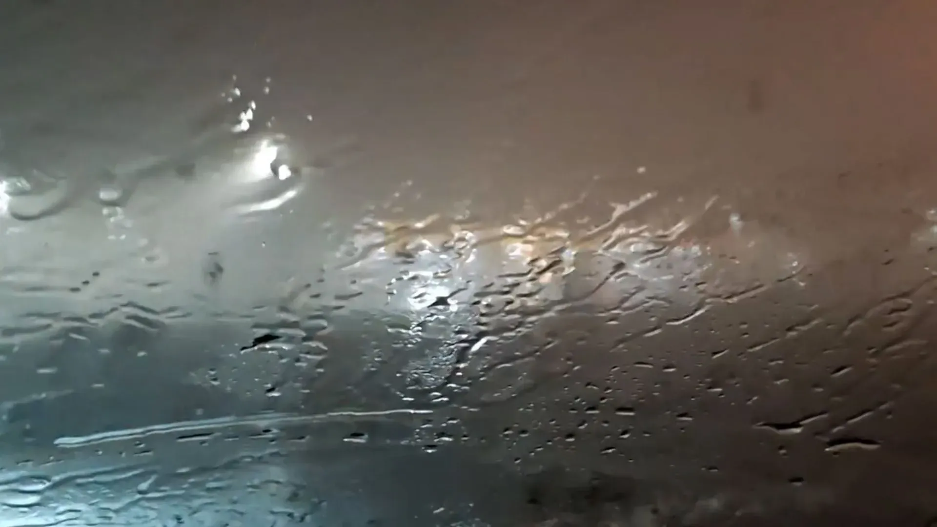مشهد لزخات المطر الغزيرة تتساقط على الزجاج، مع أضواء خافتة في الخلفية، في ظل ظروف عاصفة شديدة تهدد بحدوث أعاصير.