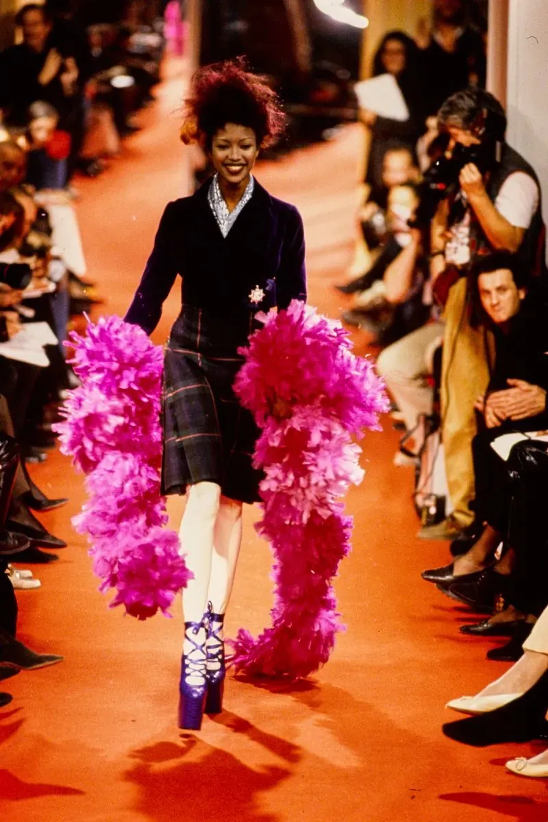 عارضة الأزياء نعومي كامبل تسير على المدرج مرتدية حذاءً عالياً وزهري اللون، مع رداء أسود وتفاصيل ملونة، أثناء عرض فيفيان ويستوود.