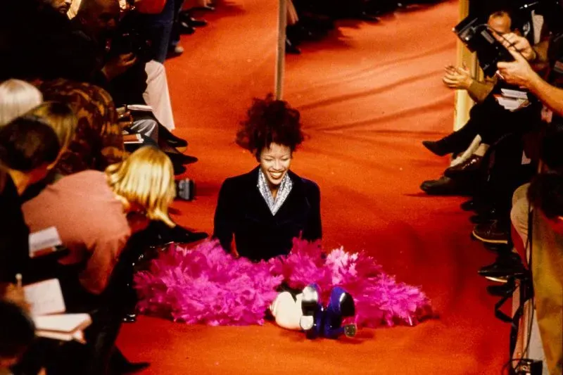 نعومي كامبل تسقط على مدرج عرض فيفيان ويستوود في باريس، محاطة بجمهور مصدوم، مع تفاصيل حذائها العالي وتنانيرها الملونة.