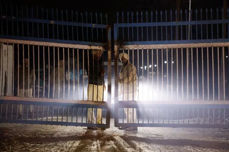 رجال أمن يفتحون بوابة معدنية في الليل، مع أضواء ساطعة خلفهم، مما يعكس مشهدًا من التوتر المرتبط بترحيل المهاجرين.