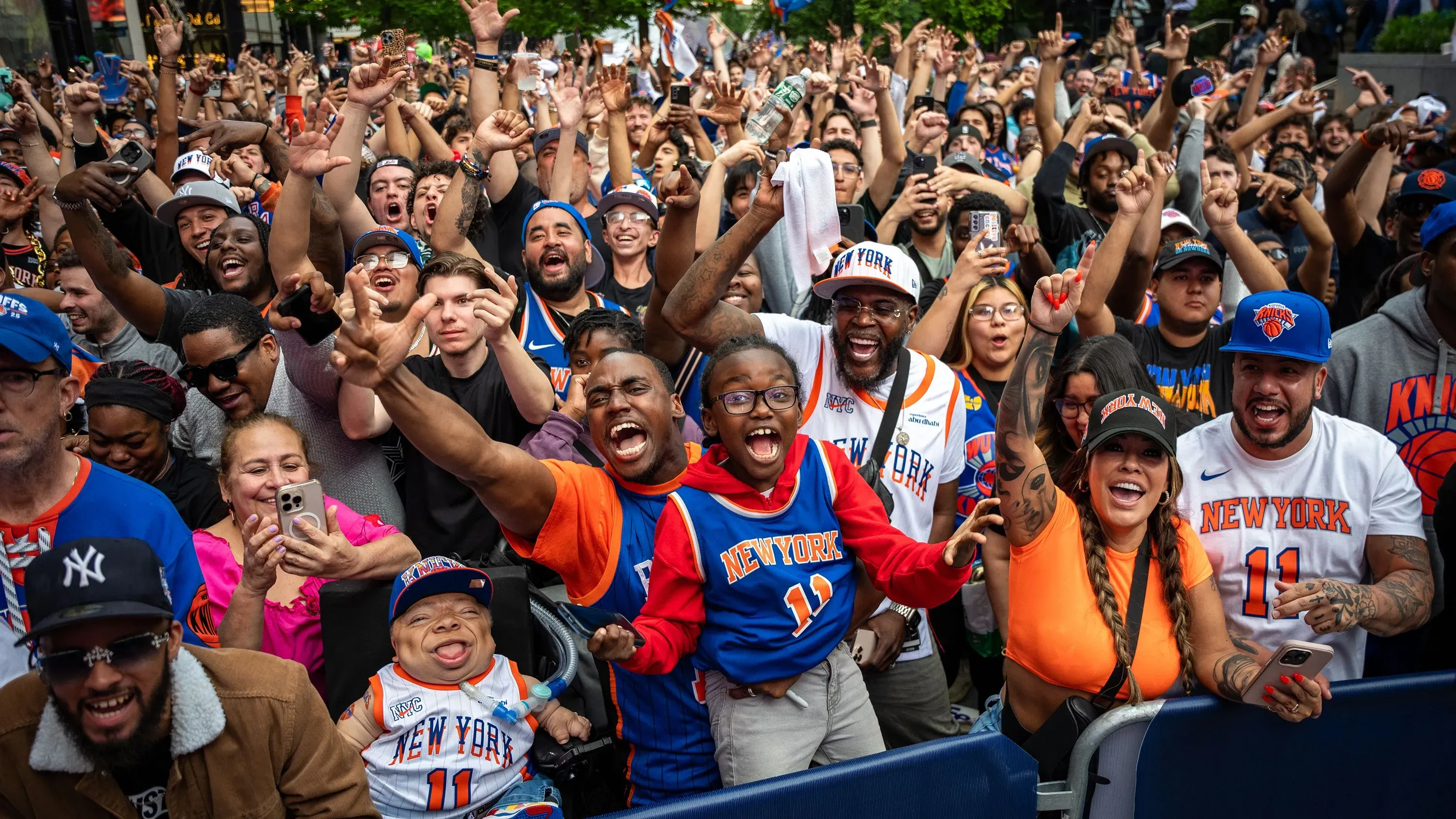 حشود من مشجعي فريق نيويورك نيكس يحتفلون في الشارع، مرتدين قمصان الفريق، مع تفاعل حماسي واضح أثناء مباريات البطولة.
