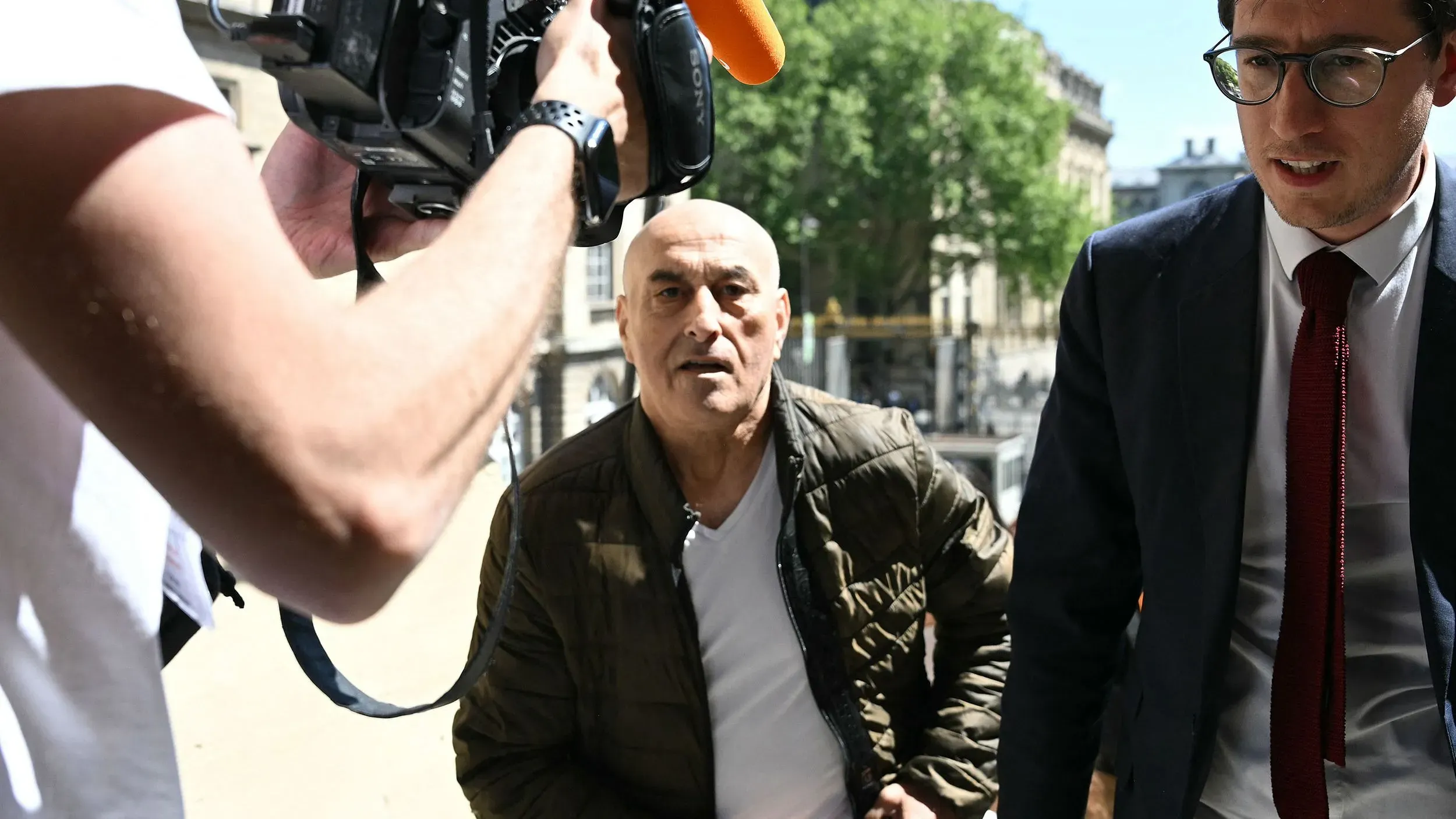 رجلان يدخلان قاعة المحكمة في باريس، أحدهما يرتدي سترة داكنة ويظهر عليه علامات الشيخوخة، بينما الآخر يرتدي بدلة رسمية.