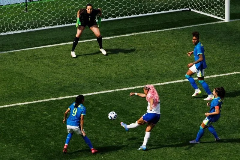 ترينيتي رودمان تسجل هدفًا في مباراة منتخب الولايات المتحدة ضد البرازيل، مع حارسة مرمى البرازيل في الخلف ولاعبات من الفريقين في الملعب.