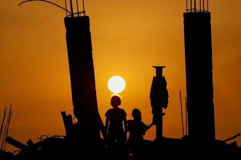 ظلال عائلة فلسطينية تتأمل غروب الشمس خلف أعمدة بناء مهدمة، تعكس مشاعر الأمل والقلق في ظل الأوضاع الحالية في غزة.