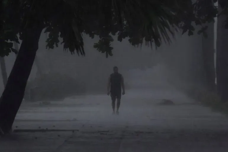 رجل يسير في شارع مبلل أثناء العاصفة الاستوائية ميليسا، مع ظروف جوية سيئة ورؤية منخفضة، مما يعكس تأثيرات العاصفة على المنطقة.