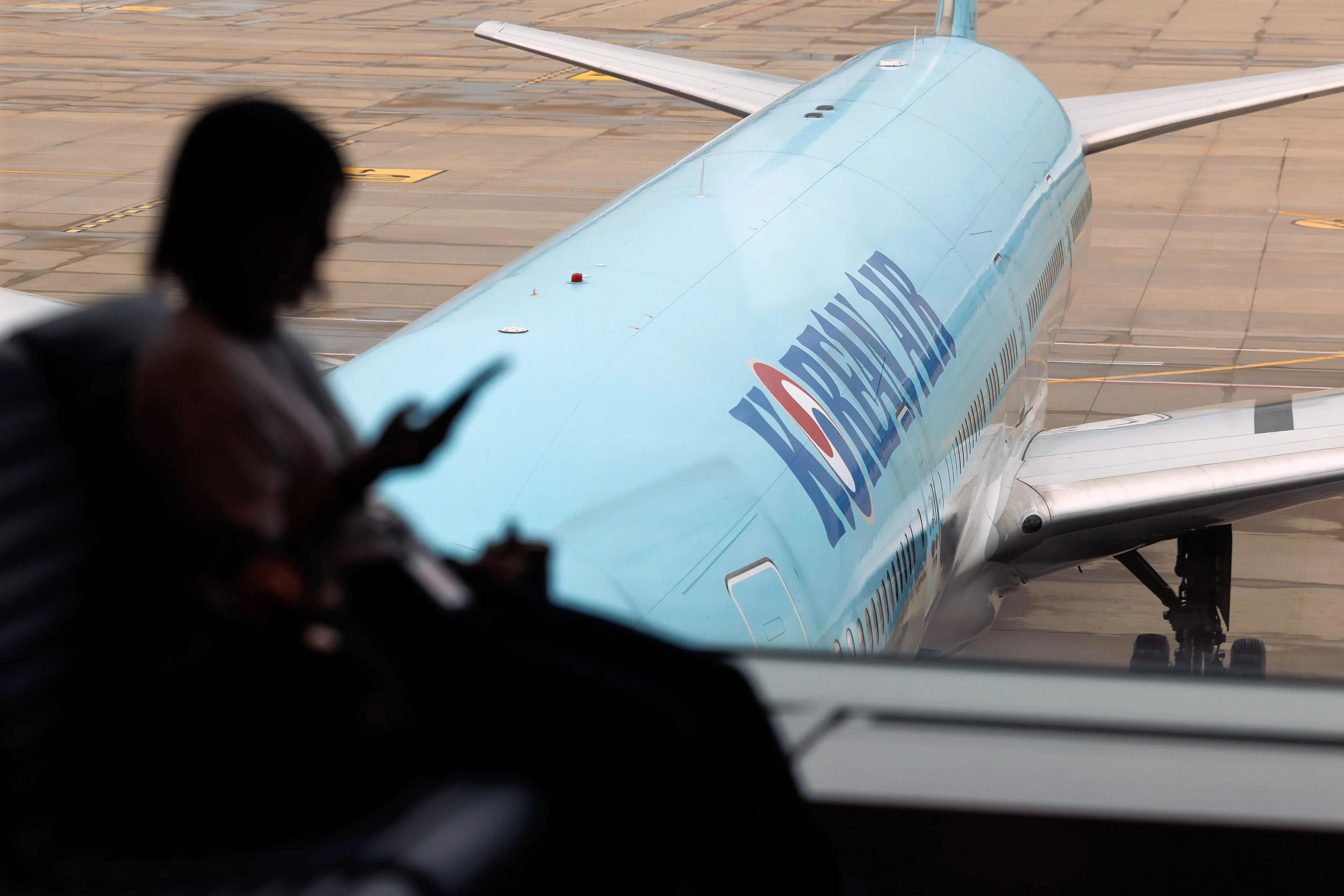 امرأة تجلس في مطار، تركز على هاتفها، بينما تظهر طائرة الخطوط الجوية الكورية في الخلفية، مما يعكس الاستثمارات الكبيرة في الطيران.