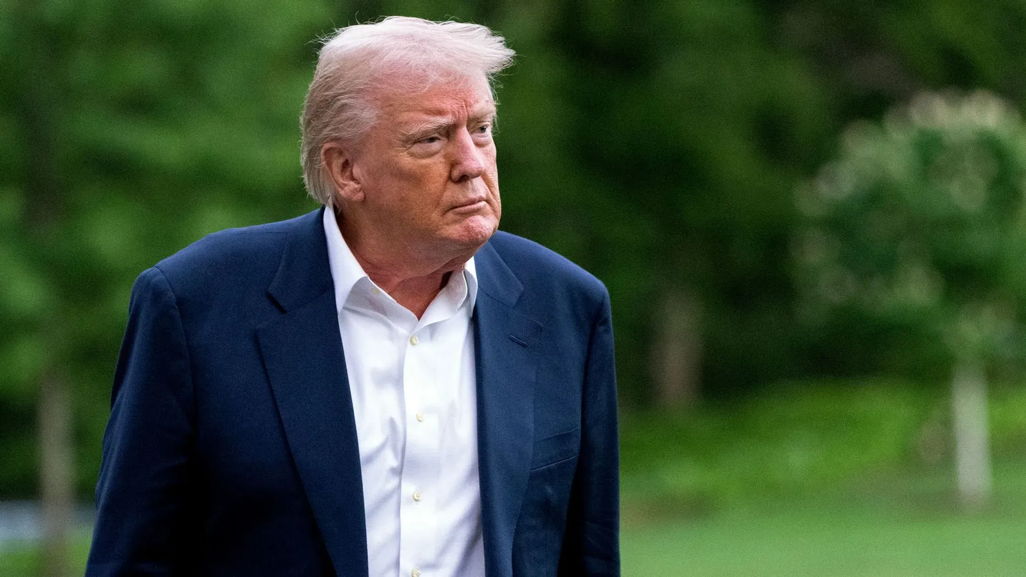 تظهر الصورة دونالد ترامب وهو يسير في الهواء الطلق، مرتديًا بدلة زرقاء، مع تعبير جاد يعكس القضايا القانونية المتعلقة بالهجرة.