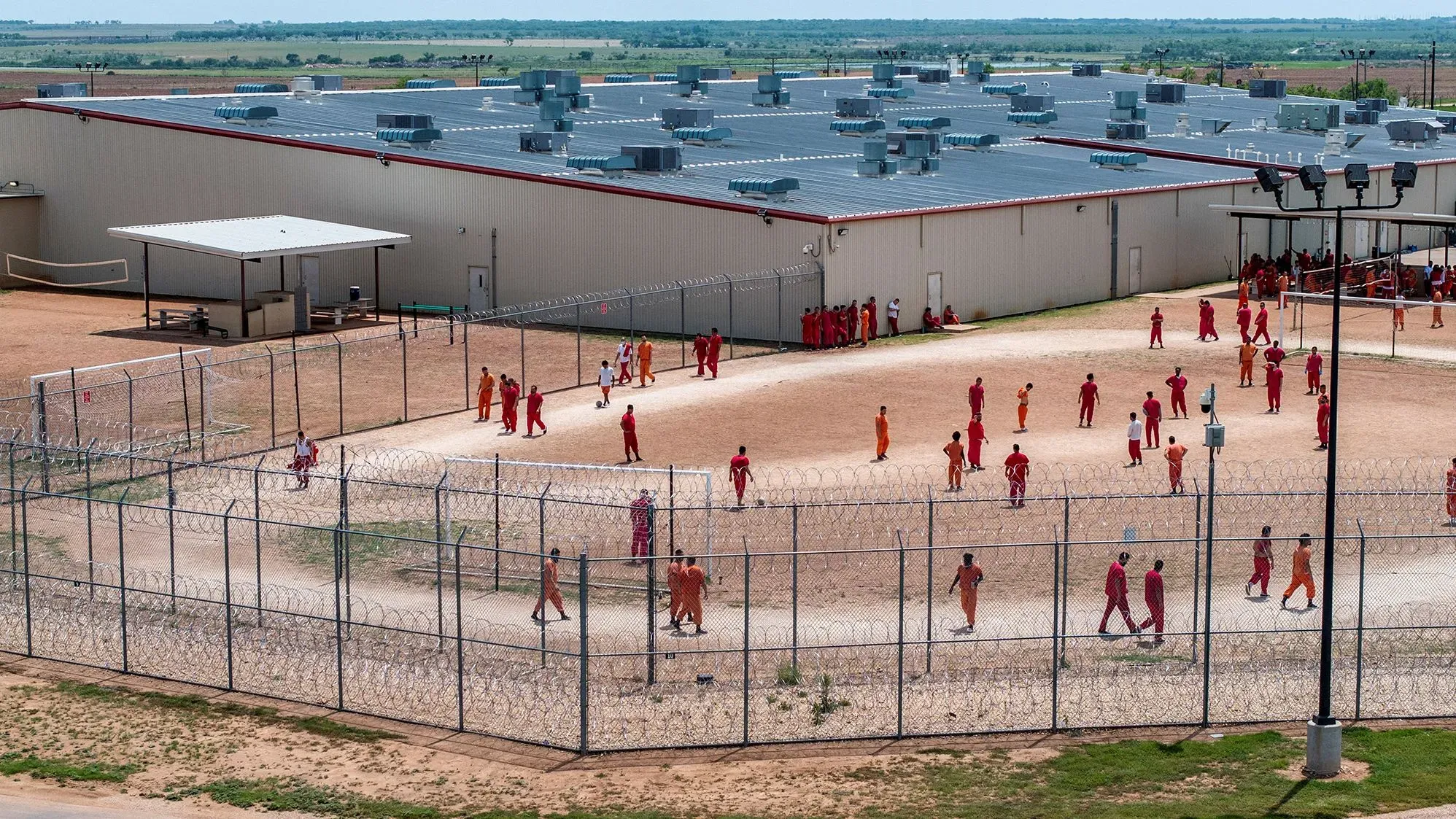 صورة لمرافق احتجاز في تكساس تُظهر مجموعة من المهاجرين يرتدون ملابس برتقالية وحمراء، محاطين بسياج شائك، في سياق قضايا الترحيل.