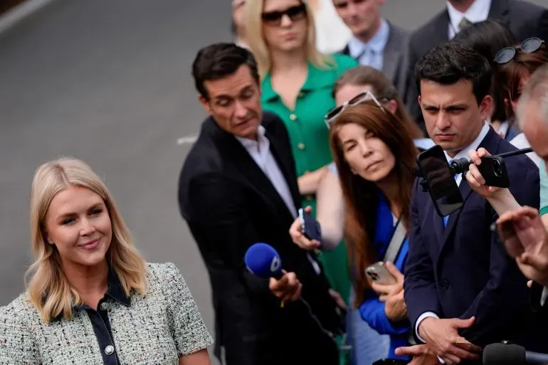 مراسلة صحفية تتحدث أمام مجموعة من الصحفيين، في خلفية مشهد يعكس التوترات حول تغطية إدارة ترامب للأخبار.