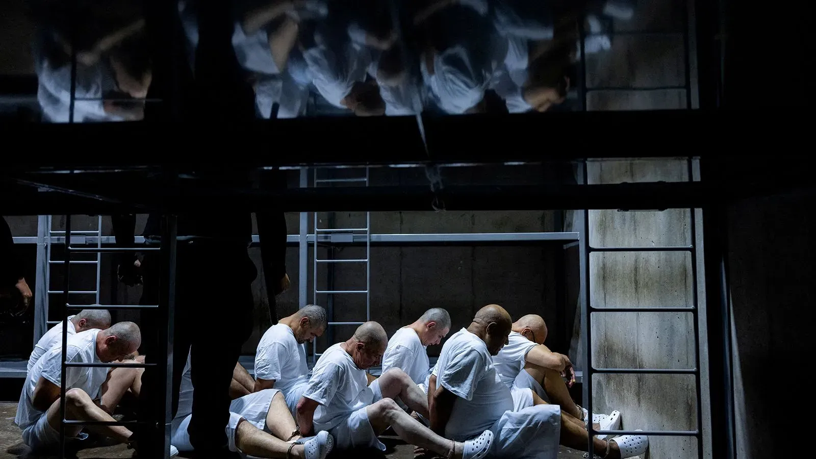 مجموعة من المعتقلين في ملابس بيضاء يجلسون معًا في مركز احتجاز مظلم، مما يعكس الظروف القاسية التي يواجهها المحتجزون في السلفادور.