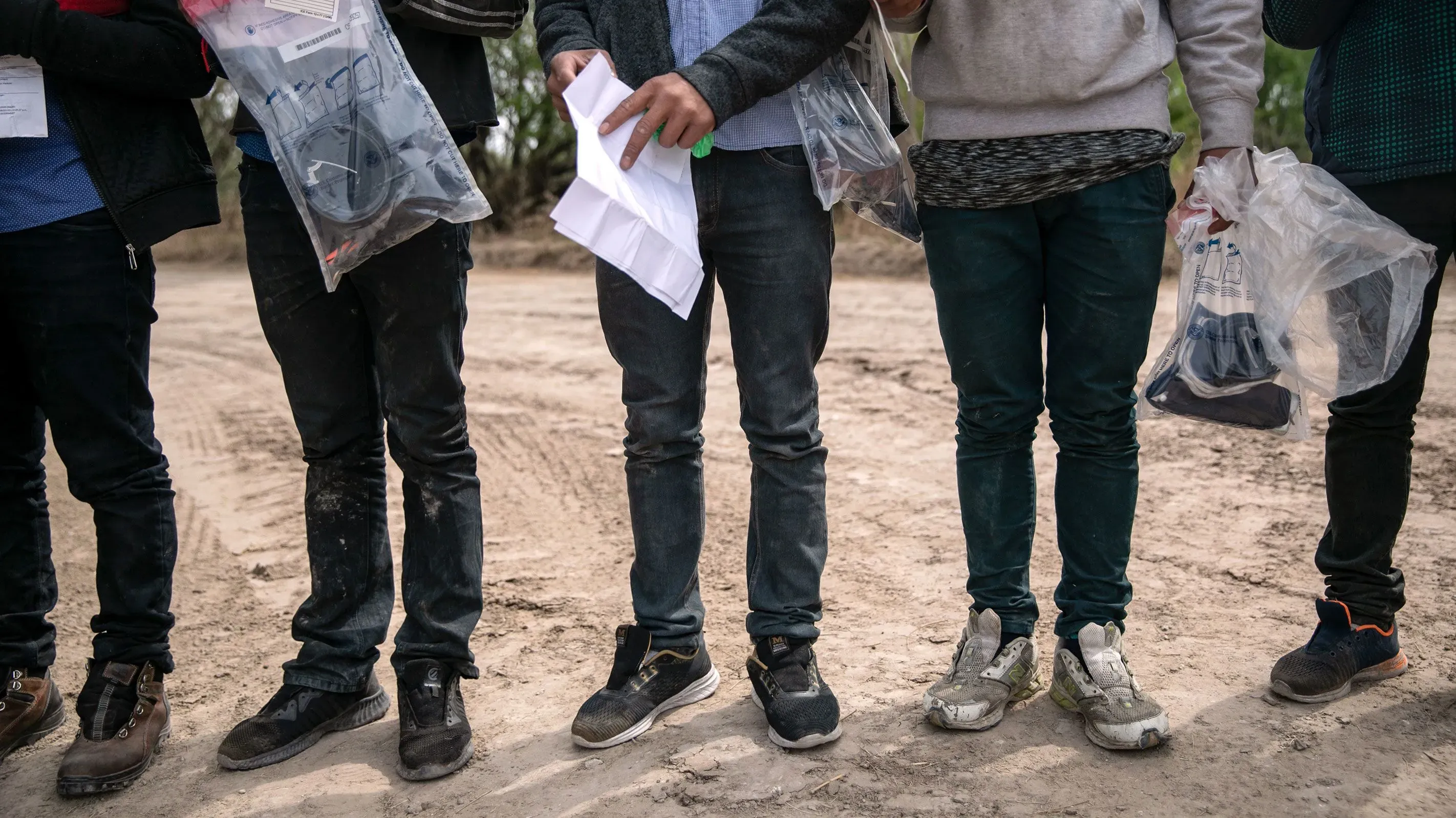 أطفال مهاجرون غير مصحوبين بذويهم يقفون في صف، يحملون أوراقًا وأغراضًا في أكياس، مما يسلط الضوء على تحدياتهم القانونية.