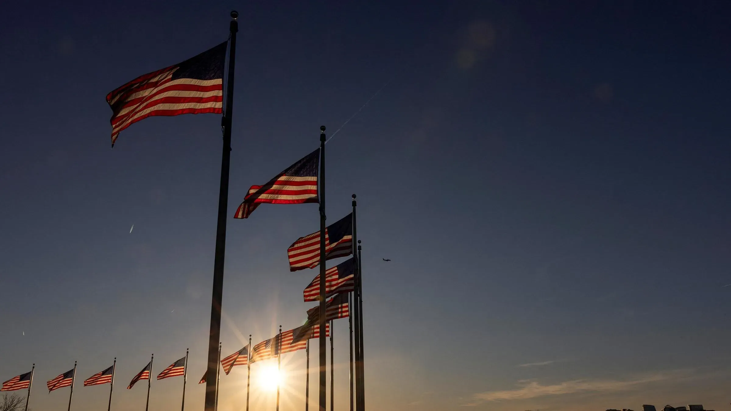 أعلام الولايات المتحدة ترفرف في الخلفية مع شمس الغروب، تعكس التغييرات المقترحة في السياسة الدبلوماسية الأمريكية.