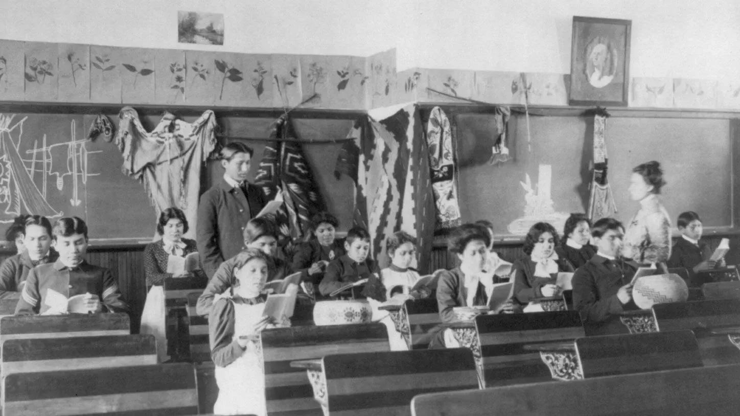 فصل دراسي قديم يظهر أطفال من السكان الأصليين في مدرسة داخلية، حيث يجلسون في صفوف مع كتب، مع معلم يتحدث إليهم.