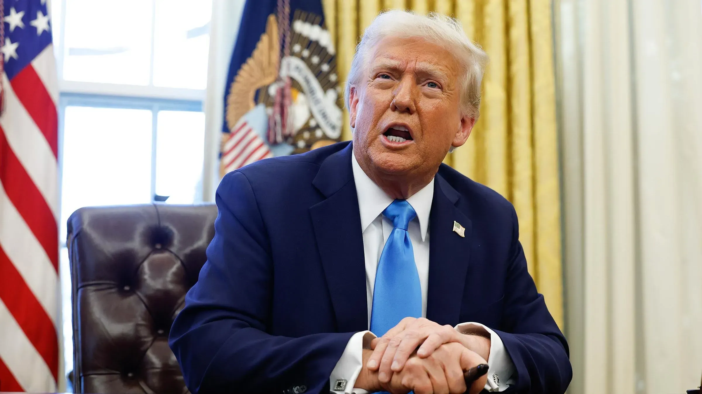 رجل يرتدي بدلة زرقاء وقميص أبيض مع ربطة عنق زرقاء، يتحدث بجدية أثناء جلوسه في مكتب رسمي، مع العلم الأمريكي خلفه.