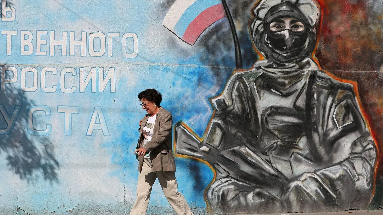امرأة تسير بجوار جدارية تصور جنديًا يحمل سلاحًا، مع العلم الروسي في الخلفية، تعكس التوترات السياسية حول أوكرانيا.