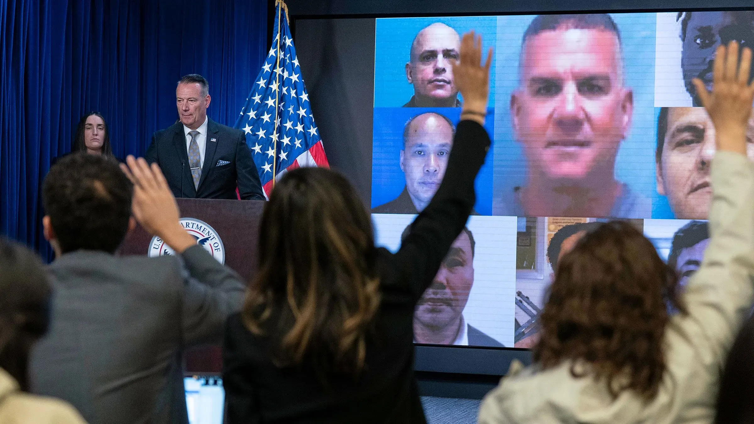 مؤتمر صحفي لوزارة الأمن الوطني حول ترحيل مهاجرين، مع صور للمحتجزين على الشاشة وخلفية تحمل العلم الأمريكي.