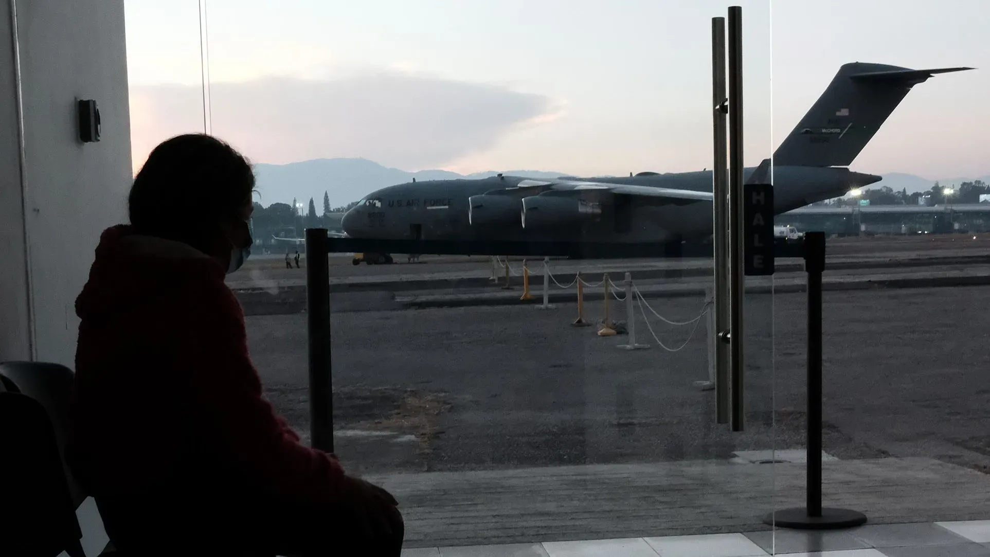 شخص يجلس في مطار، يراقب طائرة عسكرية متوقفة في الخارج، في سياق مناقشات حول ترحيل المهاجرين إلى ليبيا ورواندا.