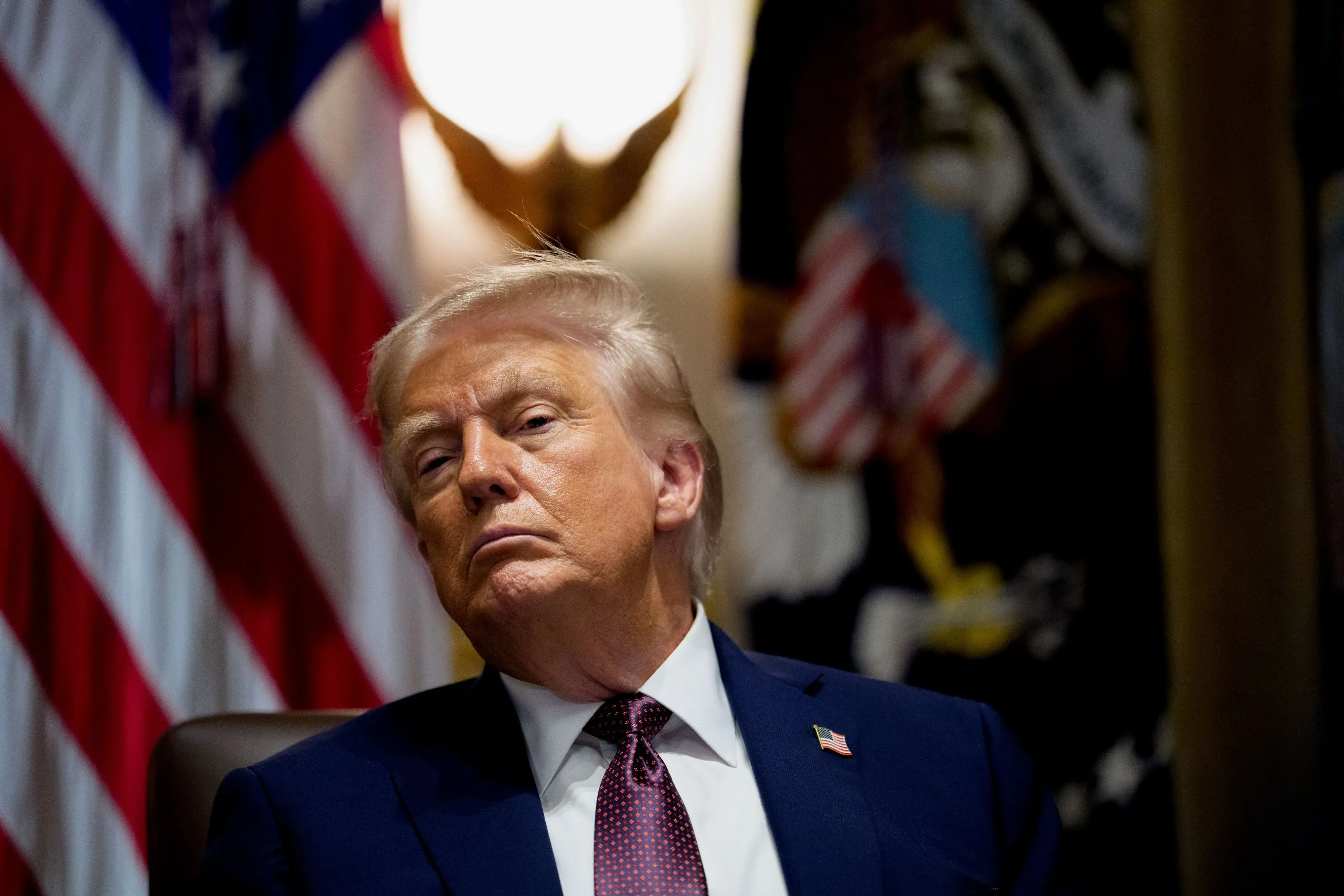 صورة لدونالد ترامب وهو يجلس في اجتماع، مع تعبير جاد على وجهه، وخلفه الأعلام الأمريكية. تعكس الصورة قوة ترامب وسلطته السياسية.