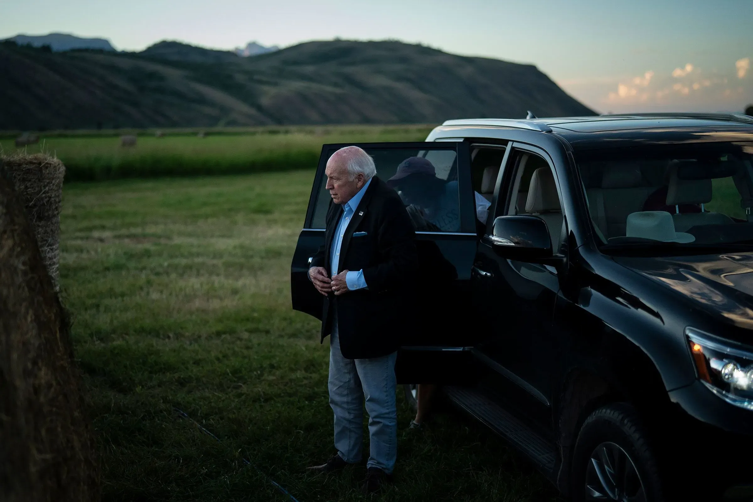 رجل مسن يرتدي بدلة داكنة يقف بجانب سيارة في منطقة مفتوحة، مع تلال خضراء في الخلفية، مما يعكس لحظة تأمل قبل جنازة ديك تشيني.