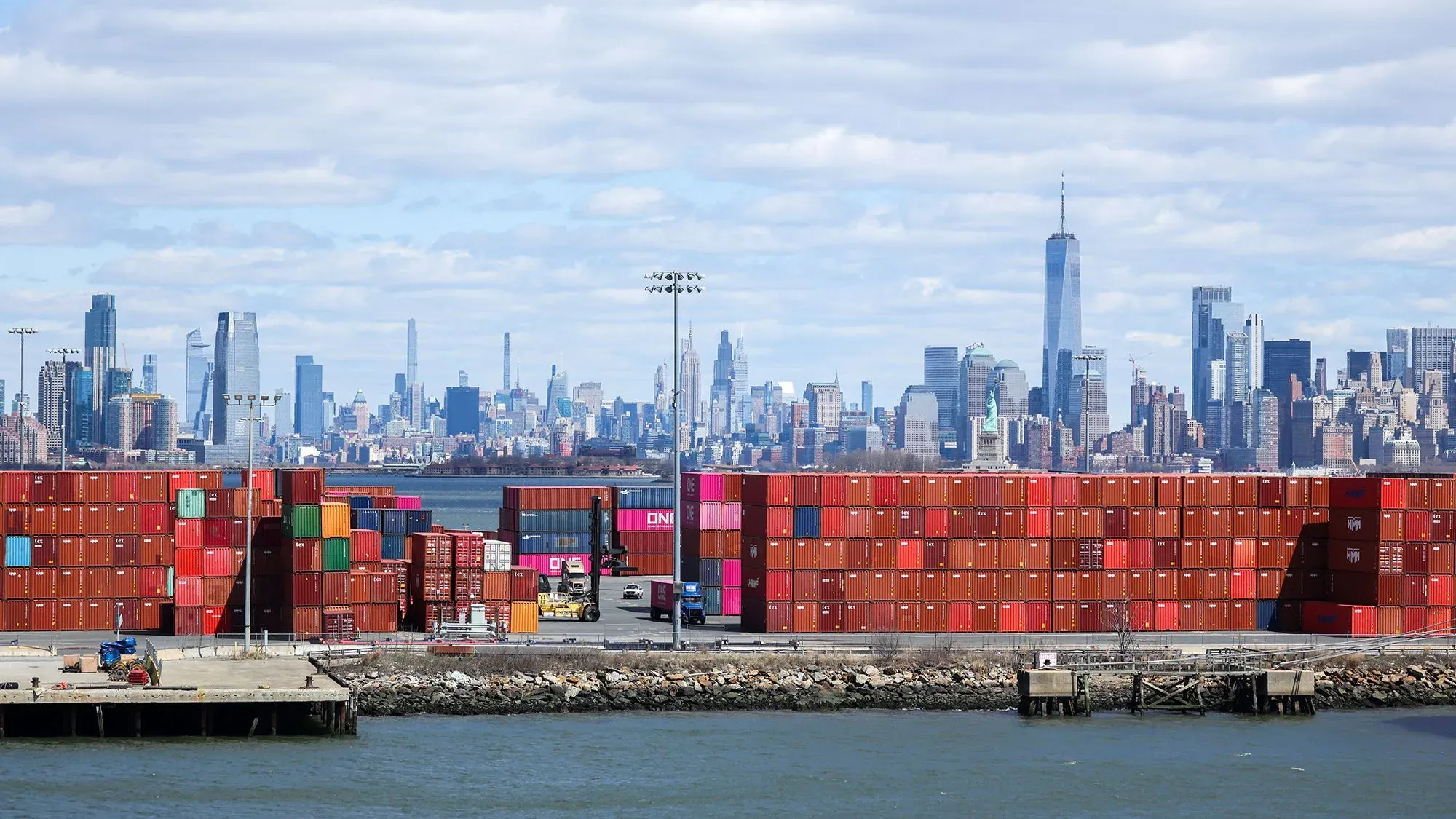 صورة لميناء يحتوي على حاويات شحن ملونة، مع skyline مدينة نيويورك في الخلفية، تعكس تأثير التعريفات الجمركية على التجارة.