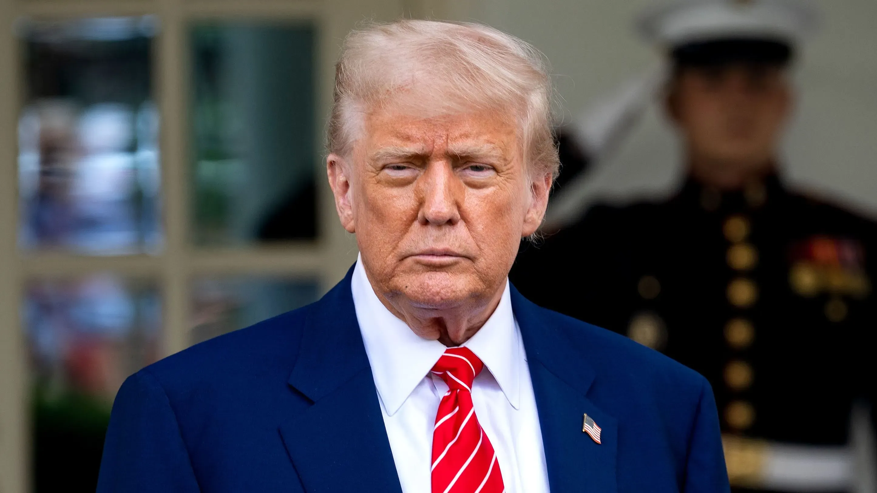 دونالد ترامب يقف أمام البيت الأبيض، مرتديًا بدلة زرقاء وقميصًا أبيض، مع ربطة عنق حمراء، في سياق حديثه عن خفض أسعار الأدوية.